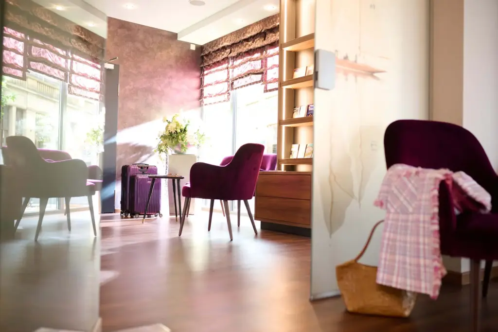 Modernes Wohnzimmer mit beigen Wänden, großen Fenstern mit roten Vorhängen, zwei lila Stühlen, einem Couchtisch mit Blumenstrauß und einem Bücherregal.